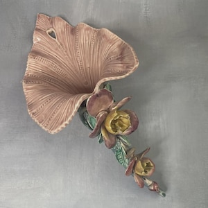 Op de afbeelding: Een vintage keramische bloemenmuursculptuur. Het heeft een groot, waaiervormig bloemblad in een gedempte roze tint, met gedetailleerde adering. Daaronder ondersteunt een stengel verschillende bloemen in paarse en gele tinten, met groene bladeren.