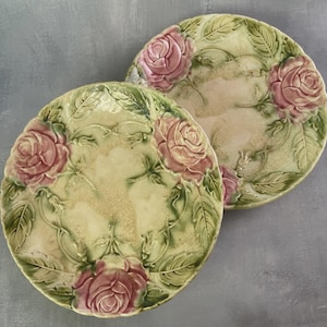 Puede incluir: Dos platos de cerámica antiguos con un diseño de rosas en relieve. Los platos presentan rosas rosas y hojas verdes sobre un fondo color crema. Los platos tienen un borde festoneado y probablemente son piezas decorativas vintage.