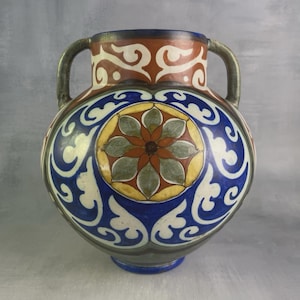 Può includere: Un vaso in ceramica con due manici, corpo arrotondato e collo largo. La superficie è decorata con un medaglione circolare centrale contenente un motivo floreale a petali multipli nei toni del marrone, verde e giallo, circondato da motivi a volute blu e bianchi.