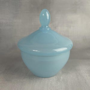 Op de afbeelding: Een lichtblauwe glazen pot met een deksel. De pot heeft een afgeronde basis en een deksel met een druppelvormige handgreep. De pot heeft een effen, ondoorzichtige kleur.