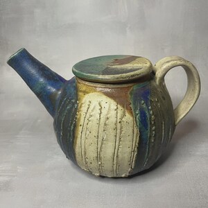 Op de afbeelding: Handgemaakte keramische theepot met een deksel. De theepot heeft een afgeronde body met een getextureerd, gebroken wit oppervlak en verticale lijnen. De tuit is blauw en het deksel is groen en bruin. De handgreep is crèmekleurig.