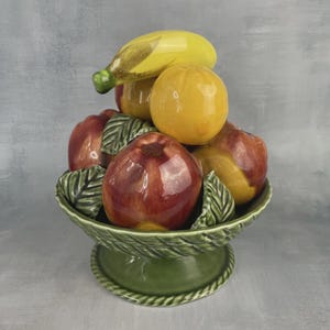 Peut inclure: Un compotier en céramique avec une variété de fruits colorés. L'arrangement comprend des pommes rouges, des bananes jaunes et des agrumes oranges, le tout dans un bol vert en forme de feuille. Le bol repose sur un socle vert.