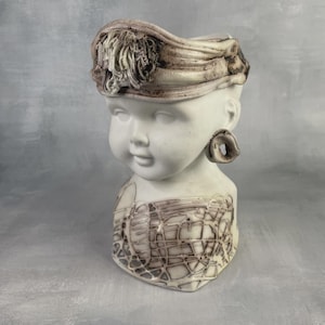 Peut inclure: Buste en céramique blanche représentant un enfant avec un bandeau et de grandes boucles d'oreilles. Le buste présente un motif détaillé sur le bandeau et l'avant. C'est un objet décoratif.