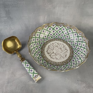 May include: A decorative serving bowl and ladle set. The bowl has a white interior with a central design and a green and purple floral pattern. The ladle has a gold-colored scoop and handle with a matching floral design.
