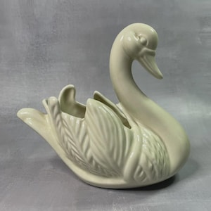 Può includere: Una fioriera a forma di cigno in ceramica color crema. Il cigno ha un collo lungo e curvo e un design dettagliato di piume sulle ali. La fioriera ha una piccola apertura per piante o oggetti decorativi. È un oggetto decorativo.