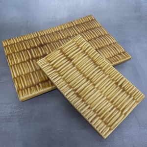 May include: Two rectangular ceramic serving trays with a textured, woven pattern in shades of tan and brown. The larger tray is behind a smaller one, both resting on a gray surface.