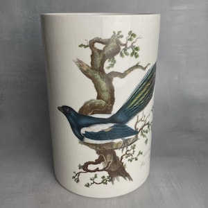 Può includere: Un vaso di ceramica bianco con una dettagliata illustrazione di una gazza appollaiata su un ramo di un albero. L'uccello ha un piumaggio nero, bianco e blu. L'albero è marrone con foglie verdi, su sfondo bianco.
