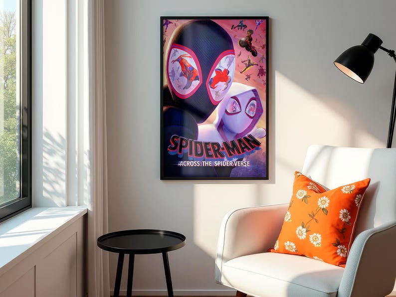 May include: A framed poster featuring Spider-Man and Spider-Gwen from "Across the Spider-Verse." The poster has a black frame and is hanging on a white wall. An orange floral pillow rests on a white armchair. A black side table is in the foreground.