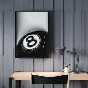May include: A framed monochrome print of a glossy eight ball, mounted on a grey wall above a wooden desk. A black desk lamp and a white mug with pens sit on the desk. The chair has a black seat.