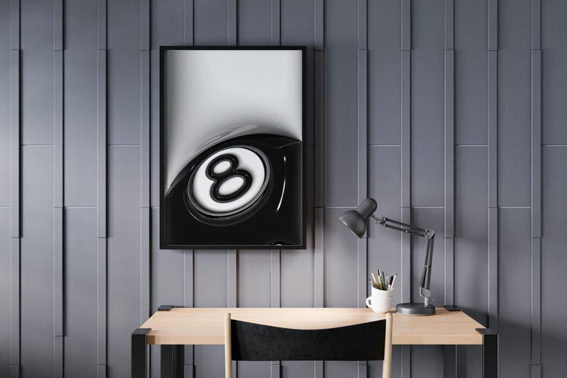 May include: A framed monochrome print of a glossy eight ball, mounted on a grey wall above a wooden desk. A black desk lamp and a white mug with pens sit on the desk. The chair has a black seat.