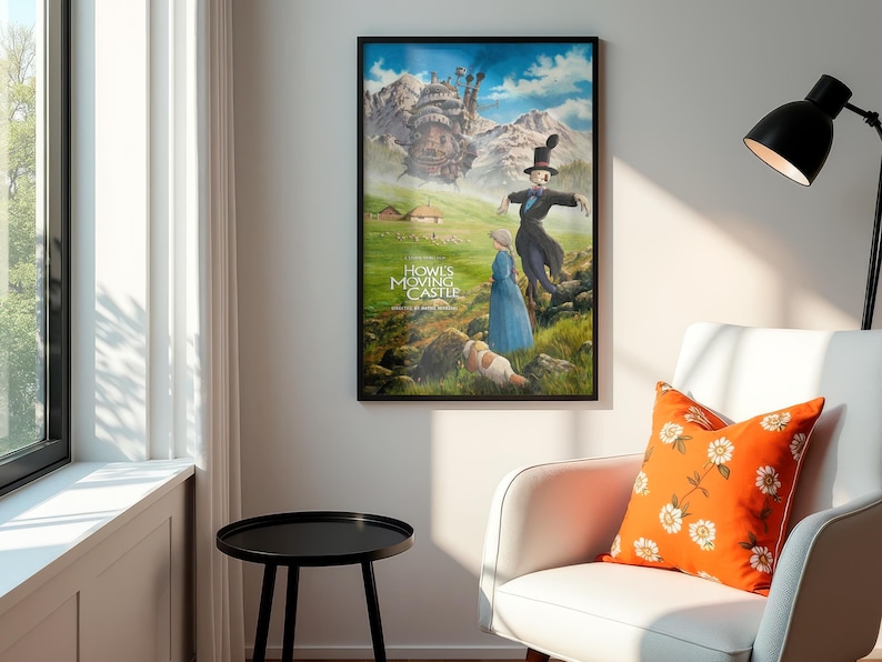Puede incluir: Un p&oacute;ster enmarcado de "El castillo ambulante" cuelga de una pared. La obra de arte representa personajes en un paisaje verde con un castillo al fondo. El p&oacute;ster est&aacute; en un marco negro. Un coj&iacute;n naranja con flores descansa sobre una silla blanca.