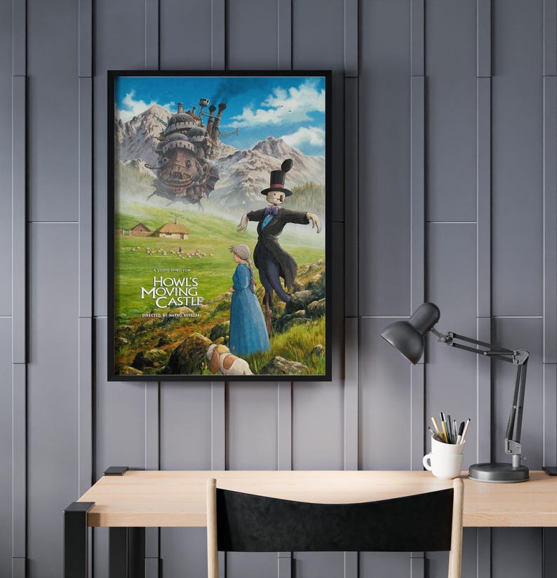 Puede incluir: P&oacute;ster enmarcado con arte de la pel&iacute;cula animada "El castillo ambulante". La imagen muestra una escena caprichosa con un castillo, personajes y texto. El p&oacute;ster se exhibe en una pared gris sobre un escritorio de madera.