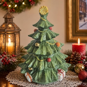 May include: A green ceramic Christmas tree figurine featuring a textured needle design, a yellow star topper, and small round ornaments in red and gold. Two white gift boxes with red and orange ribbon patterns are positioned at the base of the tree.