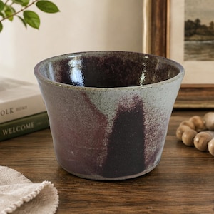 May include: A wide, tapered ceramic bowl featuring a mottled glaze with deep plum and dark charcoal tones blending into a lighter, speckled sage green exterior. The interior is finished in a dark, glossy, and reflective plum-colored glaze.