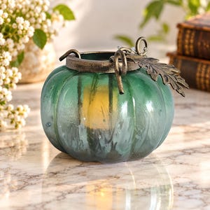 Peut inclure: Un bougeoir en verre vert sarcelle en forme de citrouille avec une texture côtelée. Le sommet présente un rebord métallique foncé avec un accent de feuille en métal décoratif et deux boucles métalliques incurvées. Une lumière jaune chaude brille.