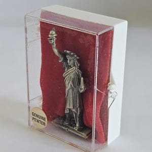May include: A pewter Statue of Liberty figurine inside a clear plastic case with a white frame. The statue is set against a red velvet background. The case has a sticker that reads "GENUINE PEWTER."