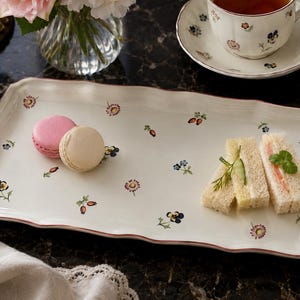 Può includere: Un vassoio da portata rettangolare in ceramica bianca con bordi smerlati e un sottile bordo rosa. La superficie presenta un motivo sparso di piccoli fiori colorati, tra cui viole e cinorrodi, con sopra due macaron e tre tramezzini.