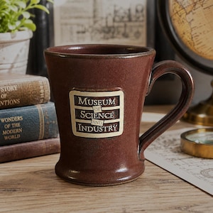 Op de afbeelding: Een donkerbruine keramische mok met een uitlopende voet en een gebogen handvat. De voorzijde van de mok is voorzien van een rechthoekig crèmekleurig embleem met de tekst "MUSEUM OF SCIENCE AND INDUSTRY" in donkerbruine, gecentreerde letters.