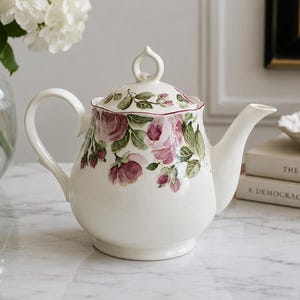 May include: A white ceramic teapot featuring a painted floral pattern of pink roses and green leaves around the upper body and lid. The teapot has a curved handle, a tapered spout, and a lid with a decorative loop-shaped handle on top.