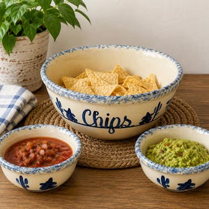 May include: A large cream-coloured ceramic bowl filled with tortilla chips, labelled with the word Chips in blue script, accompanied by two smaller matching bowls containing red salsa and green guacamole, all featuring blue sponge-painted rims.