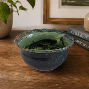 Puede incluir: Un cuenco de cerámica hecho a mano con exterior azul oscuro e interior verde. El cuenco tiene un borde ancho y una superficie texturizada. El interior presenta diseños oscuros y abstractos. Adecuado para servir o exhibir.