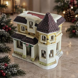 May include: A ceramic miniature house figurine featuring a cream-colored facade with yellow brick accents, a dark purple shingled roof, and a prominent turret. The structure includes multiple arched windows, a small front porch, and a wreath.