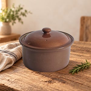 May include: A round, brown ceramic cooking pot with a matching lid. The pot features a smooth, matte finish on the sides and a glossy, darker brown glaze on the domed lid, which includes a small central knob and a tiny steam vent hole.