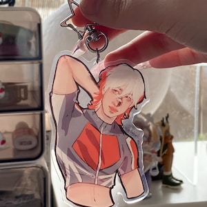 Peut inclure: Un porte-clés en acrylique transparent représentant une illustration stylisée d'une personne aux cheveux blancs et rouges, vêtue d'une tenue grise et rouge. Le porte-clés est attaché à un porte-clés en forme d'étoile argentée.