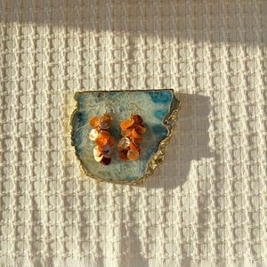 May include: A pair of dangle earrings featuring clusters of small, iridescent orange shell discs suspended from gold-toned metal hooks. The earrings are displayed on a flat, irregular piece of blue and white agate with a metallic gold-colored edge.