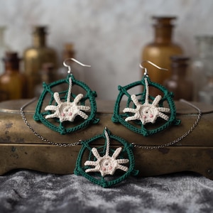 May include: A jewelry set consisting of two crochet spiderweb earrings and a matching pendant on a silver-toned chain. Each piece features a dark green circular web with a cream-colored spider in the center, accented with two small pink dots.