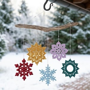 May include: A handmade mobile featuring six crocheted snowflakes in various colors: red, gold, light blue, purple, and teal. The snowflakes are suspended from a natural driftwood branch, creating a winter-themed decoration.