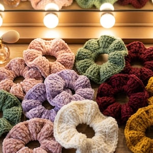 Puede incluir: Una colección de coleteros hechos a mano en varios colores, incluyendo rosa, verde, morado, burdeos y dorado. Los coleteros están hechos de un material suave y texturizado y están dispuestos sobre una superficie de madera, con un espejo de tocador en el fondo.