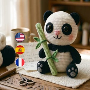 Puede incluir: Un oso panda de ganchillo hecho a mano con pelaje blanco y negro, sosteniendo un tallo de bambú verde. El panda tiene ojos negros, una nariz rosa y una pequeña sonrisa. El panda está sentado sobre una superficie de ganchillo blanca.