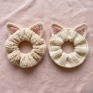 Könnte beinhalten: Zwei Haargummis in Katzenform. Einer ist ein weiches, plüschiges Beige, der andere ist cremefarben gestrickt. Beide haben rosa Innenohren. Diese Haaraccessoires sind entworfen, um Haare zu halten und eine niedliche, katzenartige Note hinzuzufügen.