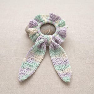 May include: A crochet hair scrunchie featuring a variegated yarn pattern in soft pastel shades of lavender, mint green, and cream. The accessory has a ruffled, circular design with two long, pointed fabric tails extending from the bottom edge.