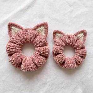 Op de afbeelding: Twee roze fluwelen scrunchies, elk in de vorm van een kattenkop, met geborduurde oren. De scrunchies zijn gemaakt van een zacht, pluche materiaal, perfect om een vleugje vrolijkheid aan elk kapsel toe te voegen. De achtergrond is een witte, getextureerde stof.
