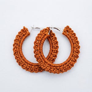 May include: A pair of hoop earrings featuring a thick, textured rust-orange crochet covering over a circular frame. The earrings include silver-toned metal posts and hinged clasps at the top, providing a secure closure for these circular accessories.