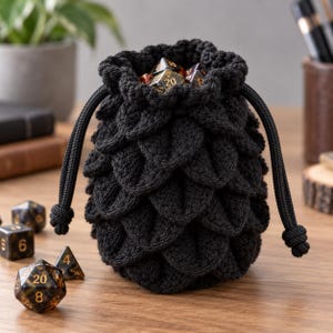 May include: A black crocheted drawstring pouch featuring a textured scale-like pattern on the exterior. The bag is filled with several multi-faceted polyhedral dice that have dark translucent bodies and gold-colored numbers etched into their surfaces.