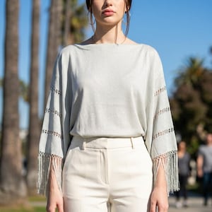 May include: A light gray poncho-style top with fringe detail on the sleeves, paired with cream-colored pants. The top has a boat neck and open sleeves with a decorative cut-out pattern. The fringe hangs from the sleeve edges.