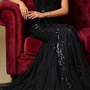 Puede incluir: Un vestido negro con cuello halter, corpi&ntilde;o con lentejuelas y falda de tul fluida. El vestido presenta una silueta de sirena y est&aacute; adornado con detalles brillantes. La modelo est&aacute; sentada en un sof&aacute; de terciopelo rojo.