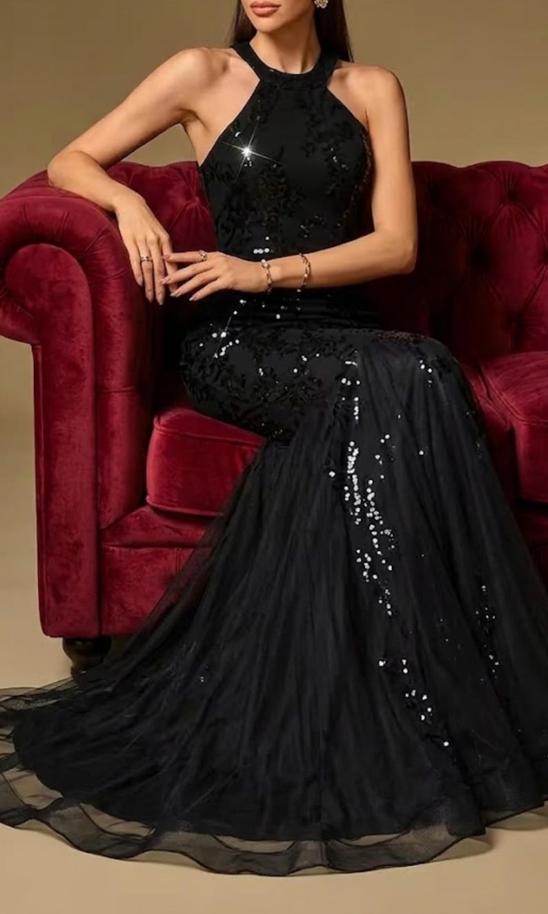 Puede incluir: Un vestido negro con cuello halter, corpi&ntilde;o con lentejuelas y falda de tul fluida. El vestido presenta una silueta de sirena y est&aacute; adornado con detalles brillantes. La modelo est&aacute; sentada en un sof&aacute; de terciopelo rojo.