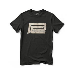以下が含まれることがあります： 黒いTシャツ。くすんだオフホワイトのグラフィックで、幾何学的なデザインで「R」と「E」の文字が描かれています。Tシャツはクルーネックと半袖です。「BAD BOYS」のブランド名がタグにプリントされています。