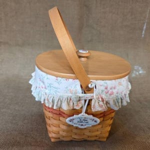 May include: A woven basket with a light brown wooden lid and a single upright handle. The basket features a ruffled fabric liner with a floral pattern in pink and green. A white tag attached to the front reads To Mom With Love.