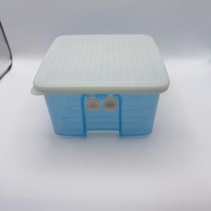 May include: A light blue plastic storage container with a white lid. The lid has a textured pattern. The container has two white latches on the front. The container is square-shaped and sits on a white surface.