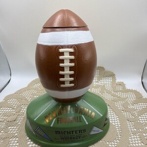 May include: A brown and white football-shaped decanter with a green base. The base is labeled "Pennsylvania Football" and "Michter's Whiskey". The decanter has white laces and stripes, and a brown lid.