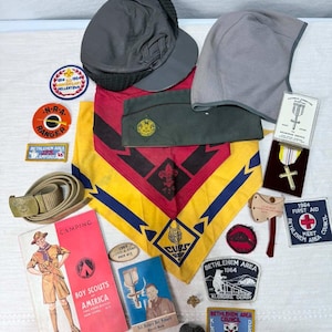 May include: A collection of Boy Scouts of America memorabilia including a grey cap, a green garrison cap, a yellow and red neckerchief, a tan belt, a religious medal, various embroidered patches, a camping handbook, and a small booklet.