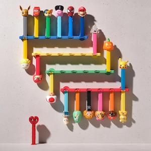 Peut inclure: Une collection de distributeurs de bonbons PEZ colorés avec des personnages de dessins animés, présentés sur des étagères murales. Les distributeurs incluent des personnages comme Pikachu et Mario, avec un distributeur en forme de cœur rouge en bas.