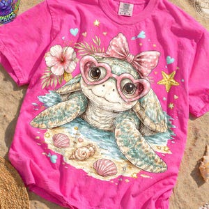 May include: A bright pink t-shirt featuring a whimsical illustration of a sea turtle wearing heart-shaped sunglasses and a bow. The design includes seashells, a starfish, and a flower, with the text "DOBSY GRAPHICS" in the upper left corner.