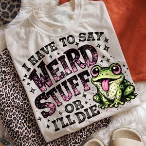 May include: Cream-colored t-shirt with the text "I HAVE TO SAY WEIRD STUFF OR I'LL DIE" and a cartoon frog graphic. The frog is green with pink eyes and tongue. The shirt is surrounded by leopard print pants and other items.
