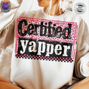 May include: Cream-colored sweatshirt with the text "Certified Yapper" in a black, distressed font. The text is set against a pink and black checkered background. The sweatshirt has a crew neck.